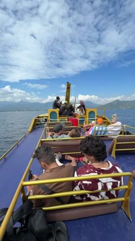 The video shows the view from the back of a yellow boat with a blue deck as it travels on the water. Several people are seated on benches on the boat. In the foreground, two men are seated facing away from the camera. One man on the left wears a brown shirt and has a black bag and flip-flops near him. The man on the right wears a burgundy and white tie-dye shirt. Further back, other passengers are visible, including a group gathered near the front of the boat around a central mast. The boat is moving towards a distant shoreline with mountains under a bright blue sky filled with white clouds. The water is calm, and the sky is clear.
