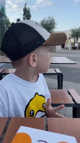 A young boy wearing a white t-shirt with a yellow cartoon shark graphic and a baseball cap sits at a table, looking to his right with his mouth slightly open. The boy appears to be in an outdoor seating area, with wooden tables and benches visible. In the background, several people are walking along a paved pathway. A couple, a man in a dark brown shirt and shorts and a woman in a light-colored floral dress and sunglasses, walk towards the camera. Further in the background, a person sits on a bench playing a stringed instrument. A large, layered mountain range with some industrial structures and trees is visible in the distance under a hazy sky. The lighting suggests it is late afternoon or early evening.