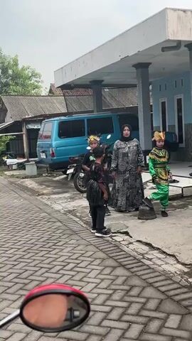 The video shows a group of people standing on a brick-paved street in front of a building with a covered walkway. A bright blue van is parked behind them, along with a motorcycle. Two children, dressed in what appear to be traditional costumes with headpieces and elaborate garments, are in the foreground. One child is wearing a black outfit with a fringe skirt, and the other is wearing a green and gold costume. An adult woman, wearing a headscarf and a patterned dress, stands near the children. The environment appears to be a residential or rural area, with simple buildings and greenery visible in the background. The lighting suggests it is daytime, possibly overcast.