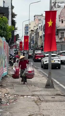 The video depicts a woman in Vietnam walking on a sidewalk next to a busy street. She wears a conical hat, a red shirt, and patterned black and white pants. A face mask covers the lower part of her face. She carries two large, red baskets suspended from a shoulder pole. The baskets appear to contain food items. The street is lined with Vietnamese flags and buildings. Cars and motorcycles fill the roadway. The sidewalk on which the woman walks is littered with trash.