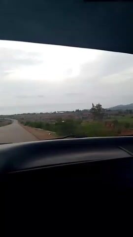 The video shows a view from inside a vehicle traveling on a road. The road curves to the left and extends into the distance. The landscape on either side of the road appears to be rural with some vegetation and sparse buildings. The sky is overcast with clouds. The foreground of the video is dominated by the dark interior of the vehicle, likely the dashboard. The footage appears to be recorded in daylight.