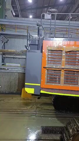 A large orange Hitachi excavator with a black track system is parked in a warehouse-like environment. The excavator has "55" and "EX1200" visible on its side. Two individuals wearing white protective suits are present. One person is standing near the excavator's arm, while the other is further back, also in a white suit. The floor appears wet and there is some water on the ground. The background includes industrial shelving and a large blue wall.