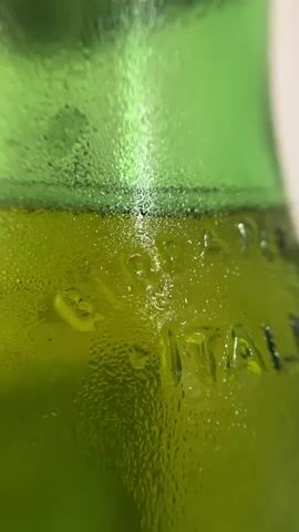 A close-up shot of a green glass bottle with condensation on its surface. Embossed lettering is visible on the bottle, with the letters "ITAL" being the most discernible. The lighting highlights the water droplets and the textured surface of the glass. The background is blurred and appears to be a light color, likely a wall or surface.
