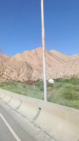 The video shows a road traveling through a mountainous landscape. The road is paved and has white lines marking the lanes. There are concrete barriers along the side of the road in some sections. The mountains are rocky and appear to be made of reddish-brown sediment. There is sparse vegetation on the hillsides, including some green bushes and grasses. The sky is clear and blue. In the distance, a white yurt can be seen among the vegetation. There are also several lampposts along the road. The perspective suggests the video is being recorded from a moving vehicle, likely a car, as it travels along the road.