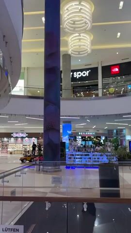The video shows the interior of a modern shopping mall. In the foreground, a glass railing overlooks a lower level of the mall. Dominating the center of the frame is a large cylindrical pillar wrapped in an LED screen displaying animated imagery. Initially, the screen shows a floral pattern, which then transitions to an animated scene with koi fish swimming among plants and rocks. Further back, other pillars with LED screens are visible, displaying different animated visuals, including one with a blue, wave-like pattern and another with a tropical beach scene. In the background, various retail stores are identifiable, including "spor," "The North Face," and "Salomon." People are seen walking and shopping on different levels of the mall. The mall features modern architecture with high ceilings, circular light fixtures, and multiple levels connected by escalators and walkways. The overall impression is a bright, spacious, and technologically advanced shopping environment.