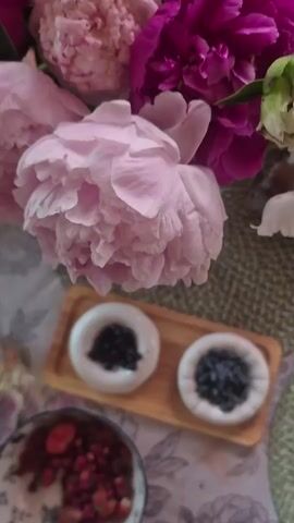 This is a close-up video of a table setting with a floral arrangement. The main subjects are two white ceramic dishes on a wooden tray, each filled with different types of berries. The left dish contains dark, round berries, possibly blueberries or blackcurrants, while the right dish holds elongated, bluish-purple berries, likely honeysuckle berries. In the foreground, a bowl overflows with small strawberries and other red berries, and a hand holding a spoon is partially visible, suggesting someone is about to eat them.

The background is dominated by a lush bouquet of peonies in shades of pink and magenta, with some greenery. The table is covered with a patterned tablecloth featuring floral motifs. A textured placemat is visible beneath the wooden tray. The lighting suggests it's daytime, with soft light illuminating the scene. The overall atmosphere is serene and natural, evoking a sense of abundance and the simple pleasure of fresh fruit. No text is visible in the video. The location context is Астра-1 Днт, Russia.