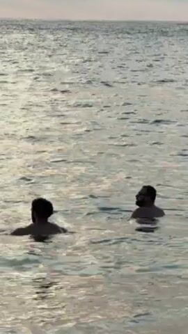 Two men are in the ocean, waist-deep. One is facing away from the camera, and the other is facing the camera. The water is shimmering in the sunlight. The horizon is visible in the background.