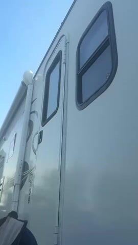 The video shows a side view of a white RV. The RV has a closed door with a window and a second window above it. An awning is partially extended on the side of the RV. People are visible in the foreground, partially obscured. One person is wearing a red shirt and a hat. Another person is wearing sunglasses and a grey hat. A blue umbrella is also visible in the foreground, angled upwards. The background is a clear blue sky with some trees and other RVs in the distance. The overall environment appears to be a campground or outdoor recreational area.