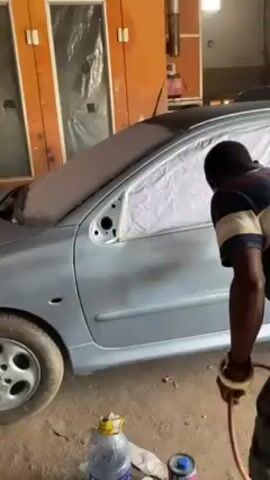 In a medium shot taken inside what appears to be an auto body shop, a man is spray-painting the side of a light blue car. The car's windows and parts of its body are covered with paper or plastic to protect them from the paint. The man is holding a spray gun connected to a hose. He is wearing a striped shirt and is focused on evenly coating the car's surface with paint. In the background, there are cans of paint and other shop equipment. The shop itself seems to be an indoor space with light-colored walls and a few windows, providing natural light in addition to the overhead lighting.