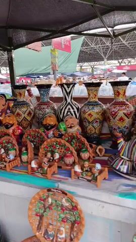 A stall at an outdoor market displays a variety of ceramic figurines and decorative vases. The figurines are of people, many with exaggerated features like mustaches and colorful clothing. There are also smaller ceramic houses with yellow accents. The vases are tall and ornate, decorated with intricate patterns. Some are displayed behind the figurines, while others are further back on the stall. In the foreground, several large, decorative plates with detailed patterns are visible. The stall is covered by a dark canopy, and beyond it, more market stalls and structures can be seen. There are no discernible people actively interacting with the merchandise in this shot.