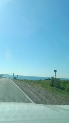 The video shows a view from the windshield of a car driving on a road. The road is asphalt and curves to the left. On the right side of the road is a grassy embankment, and beyond that, a large body of water, likely a sea or large lake, stretches to the horizon under a clear blue sky. There are a few trees on the left side of the road. In the distance, there are some cars on the road, and a construction crane is visible. A traffic sign, which appears to be a red circle with a diagonal line, is on the right side of the road. Another white cross-shaped monument is visible further down the embankment on the right. The sun is shining brightly.