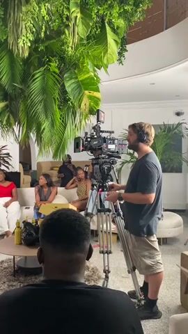 A man wearing headphones and khaki shorts operates a professional video camera mounted on a tripod. He is looking to his left, towards a group of people seated on a white couch. The camera is positioned in the foreground, obscuring the lower half of a person with dark skin and a black shirt, who is facing away from the camera. Behind the camera operator, other individuals are visible. To the left, three women are seated on a couch. One woman, wearing a red top and white pants, smiles. Another woman wears a gray top, and a third wears a patterned top. In the background, a man wearing a black shirt and a cap stands. The environment appears to be an indoor space with large, lush green plants hanging from the ceiling and potted plants in the background. There is a coffee table in front of the couch with several yellow boxes and a black bag on it. To the right of the camera operator, another person wearing headphones and a black shirt faces the camera. The scene suggests a film or video shoot is in progress.