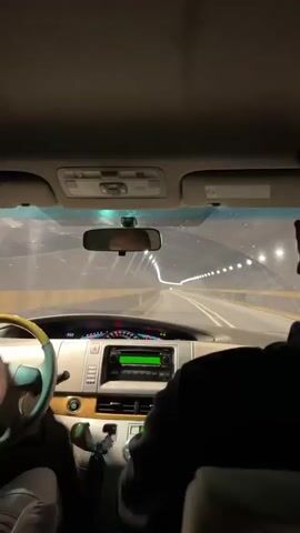 The view from the driver's seat of a car as it drives through a tunnel. The car is gray and the interior is beige. The dashboard is visible with the speedometer and radio display. The driver's hands are on the steering wheel and another person is in the passenger seat. The rearview mirror reflects the driver's face. The tunnel is brightly lit with rows of lights and has a yellow barrier on either side of the road. The road is gray and has white lane markings.