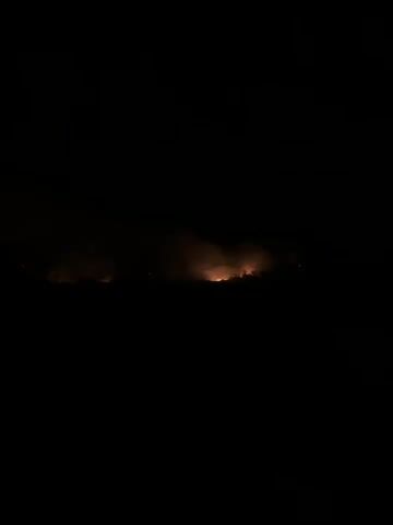 The video shows a nighttime landscape dominated by darkness. A visible horizon line is marked by a faint orange glow, interspersed with plumes of smoke. The glow suggests an active fire or multiple light sources in the distance. The smoke is illuminated by the same orange light, creating a hazy effect against the black sky. No people or specific objects are discernible due to the low light conditions. The environment appears to be rural or undeveloped, with the glow emanating from a low-lying area.