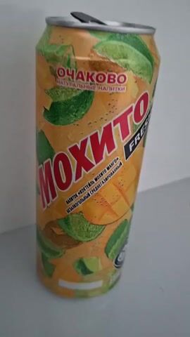 A close-up shot focuses on a single, unopened can of "MOJITO FRESH MANGO" beverage, presented against a neutral, lightly colored background. The can, primarily orange and yellow, is adorned with illustrations of mango slices, lime wedges, and mint leaves, suggesting the drink's flavor profile. Prominently displayed is the brand name "OCHAKOVO" and the product name "MOJITO" in large, bold, red lettering, with "FRESH" in smaller, black text below. A circular halal certification symbol is visible near the bottom of the can. The can is positioned upright, with its silver pull-tab slightly ajar, indicating it has not been fully opened. The lighting is soft, casting a gentle shadow to the left of the can. The overall scene is static, with no visible movement or action.