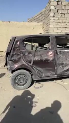 The video shows the aftermath of what appears to be a vehicle explosion in a desert-like area. There are two extensively damaged cars. The first is a maroon-colored hatchback with significant damage to its sides, missing windows, and visible interior. The second vehicle, likely a larger SUV, is black and even more heavily damaged, with its roof caved in and front completely destroyed. The surrounding environment features dilapidated brick structures or ruins, suggesting a conflict zone or area of significant destruction. Several men are gathered near the second vehicle, some dressed in traditional Afghan clothing (shalwar kameez and pakol hats), observing the scene.  The day is sunny with clear skies. The overall scene suggests a tragic event involving significant destruction and loss.
