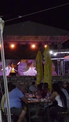 The video shows a nighttime outdoor event. A stage is set up with performers dancing. The performers wear ornate costumes with sparkling lights. The stage has a banner across the top with text. In front of the stage, an audience is seated at tables, some of whom are eating and drinking. Yellow umbrellas are positioned behind the audience. The overall atmosphere appears to be festive.