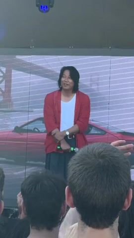 The video captures a person on a stage, likely during an event. The subject is a man with long, dark hair, wearing a white t-shirt, a red cardigan, and dark pants. He is holding a microphone in his right hand and appears to be speaking or interacting with an audience. The backdrop is a large screen displaying an image of a red car and a bridge, possibly a reference to the Fast & Furious franchise, given the subject's resemblance to Sung Kang, who plays the character Han Lue. In the foreground, the backs of people in the audience are visible, with some hands raised. The lighting suggests it is daytime or a well-lit indoor or outdoor venue. The man smiles and bows at one point, indicating a positive and appreciative atmosphere. The context provided states the location is Cesena, Italy, suggesting this is a public appearance or promotional event in that city.