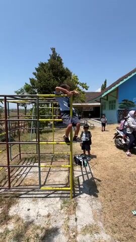 The video shows a playground scene with several children. In the foreground, a young boy wearing a blue shirt and dark shorts is climbing a yellow and silver monkey bar structure. His left leg is on a horizontal bar, and his right leg is bent upwards, with his foot near a vertical support. His arms are gripping the structure. To his right, another boy in a striped shirt and dark shorts is standing and looking at the climber, with a black bag on the ground near him. In the background, a blue and green building, possibly a school, is visible. Other children are present in the background, one of whom is walking away from the building. The ground is covered with dry grass and some concrete paths. The sky is clear and blue, indicating a sunny day. The overall setting appears to be an outdoor play area during daylight hours.