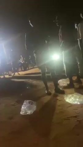 At night, a group of people are gathered outdoors. There is a bright light source to the left, creating long shadows. The ground appears to be dirt or sand. Some people are standing, while others are seated in the background. There is trash on the ground. People are moving around and interacting, but their specific actions are not clear. The overall scene appears somewhat chaotic and dimly lit.