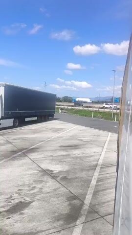A parked truck with a dark blue trailer is visible on the left side of the frame. In the background, a highway with several vehicles, including another truck and a car, is seen under a blue sky with white clouds. The scene appears to be a rest stop or truck stop with a concrete surface marked with white lines.