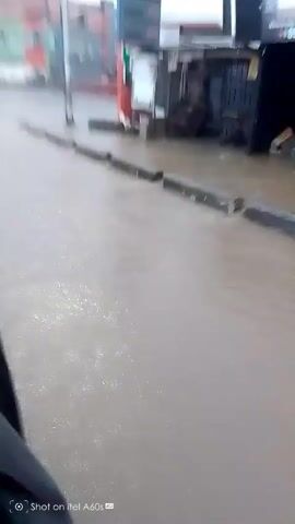 The video shows a flooded street. Water covers the asphalt, reaching up to the curbs which are visible as raised concrete barriers. In the background, there are buildings, including what appears to be a gas station with red and white pumps. A tanker truck is also visible, partially submerged in the floodwaters. The sky is overcast, suggesting rainy weather. The camera is moving, likely from within a vehicle, as a dark object, possibly the side of the vehicle, is in the foreground on the left. There are no people visible in the frame. The overall impression is a scene of moderate flooding in an urban or semi-urban area.