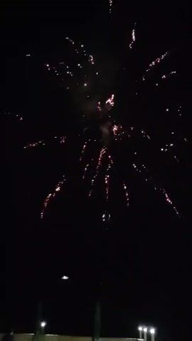 The video captures a nighttime fireworks display. The fireworks are predominantly orange and yellow, creating a starburst effect against the dark sky. Some fireworks also have trails of white and purple sparks. In the foreground, the top of a dark structure, possibly a building or a stage, is visible, along with some green objects that could be umbrellas or tents, and a few lights. There are also faint silhouettes of people at the bottom left of the frame. The overall atmosphere suggests a celebratory event.