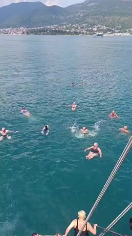 The video captures a sunny day at sea, likely in the afternoon, with clear skies and calm waters. A group of people are swimming and enjoying themselves in the turquoise sea. Some are floating on their backs, others are swimming with visible strokes, and one person is splashing in the water. A boat is partially visible in the foreground, suggesting the people are either disembarking from or preparing to board. In the background, a scenic coastline with buildings and mountains hints at a location like Đuraševići, Montenegro, with several yachts anchored offshore. The overall mood appears relaxed and joyful, indicative of a leisure activity on a pleasant summer day. No visible text is present in the video.