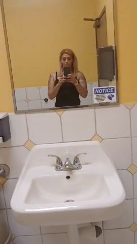 The video shows a restroom interior. The walls are painted yellow with a white tile border featuring yellow diamond accents. The restroom includes a white porcelain sink with chrome fixtures, a toilet with a gray soap dispenser mounted above, and metal grab bars alongside the toilet. Two framed paintings adorn the walls: one depicting a European-style street scene with a cafe, and the other featuring an image of three orange flowers. A black tissue dispenser is mounted on the wall above the tile border. The reflection of a woman is visible in a mirror above the sink. She is holding a smartphone, possibly taking the video.