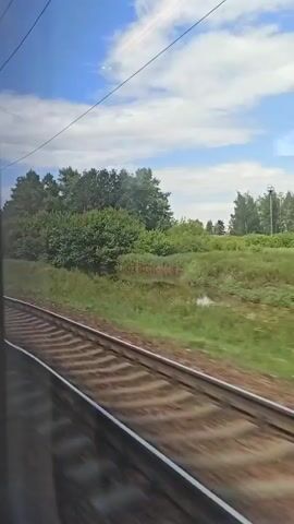 The video shows a view from a moving train looking out the window at a rural landscape. Train tracks are visible in the foreground, curving to the left. Beyond the tracks, there is a field of green grass and trees. A tall utility pole with wires is also visible. In the distance, a large, light-colored industrial building with a fire escape is seen. The sky is blue with scattered white clouds. The overall impression is of a journey through the countryside on a sunny day.