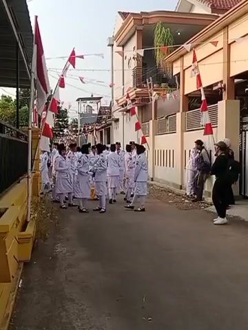 The video shows a street scene with several people, many of whom are wearing white uniforms. They are gathered in what appears to be a residential area, with houses visible on either side of the street. Indonesian flags and red and white triangular pennants are strung across the street, suggesting a national celebration or event. Some individuals in the foreground are dressed in black, and at least one person appears to be filming the group. The people in white uniforms are arranged in a somewhat formal manner, with some standing in lines and others in smaller groups. The overall atmosphere seems to be one of preparation or anticipation for an event.