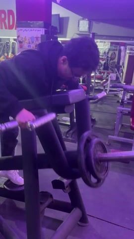 A man wearing a black hoodie, black pants, white sneakers, and glasses is performing a weightlifting exercise in a gym. He is using a machine that supports his torso while he bends over. He is holding a barbell with weights on either side, lifting and lowering it. The gym is filled with various exercise machines, and another person is walking in the background. The lighting in the gym is a mix of white and purple.