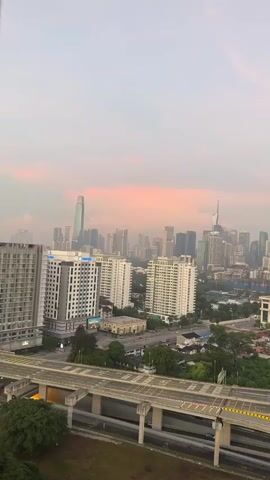 A vertical, slow-panning video shows an aerial view of a city skyline during twilight. In the foreground, a multi-level highway or overpass stretches across the frame, with concrete supports and yellow striped railings. The highway is empty of traffic. Beyond the highway, several modern apartment buildings are visible, characterized by white facades and numerous windows. Further in the distance, a dense collection of skyscrapers forms the city's core. The sky is a soft, hazy pink and orange, suggesting either sunrise or sunset, with a few wispy clouds. The overall atmosphere is tranquil and urban.