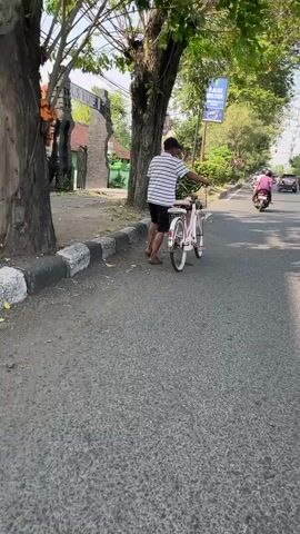 The video shows a man with dark skin and short hair, wearing a white and blue striped t-shirt, black shorts, and a black baseball cap, pushing a pink bicycle with white tires and pink handlebars. He is on the side of a paved road, next to a black and white curb. Behind him is a tree with green leaves. To the left, there is a building with a stone gate and a green wall. To the right, there is a sign that says "POLISI MILITER" in blue letters. Further down the road, a person on a pink motorcycle is visible. The environment appears to be a sunny outdoor setting with trees and buildings.