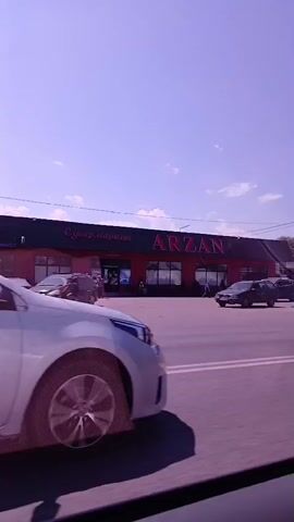 A white car drives past several businesses under a clear sky. The businesses include one with a red sign that says "ARZAN," another with a white sign that appears to say "КУЛЬТУРНЫЕ ДРУ" and "СТРОИТЕЛЬНЫЙ МАГАЗИН," and a smaller red structure with "TABAK" in white letters. Other cars and vans are parked or driving in the vicinity.