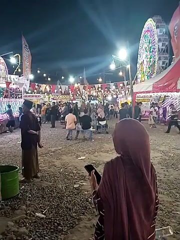 The video shows an outdoor night market or fairground scene in Sragen, Indonesia, under a dark sky. Numerous people, including adults and children, are present. In the foreground, a woman wearing a maroon hijab stands with her back to the camera, holding a mobile phone. Nearby, a man in a dark shirt and a sarong-like garment stands beside a green bucket.

The fair features various attractions, including a brightly lit Ferris wheel in the background and a small carnival train or roller coaster ride with colorful lights, which people are riding or watching. Several tents, one distinctly red and white striped, are set up. The area is illuminated by overhead string lights, streetlights, and colorful lights on the rides and tents, creating a festive atmosphere. Bunting is strung between poles. The ground appears to be unpaved, possibly dirt or gravel. People are seen walking, sitting on benches, and enjoying the lively event.