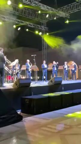 A band performs on a stage, illuminated by bright yellow spotlights. The band includes several musicians playing brass instruments and saxophone players, all wearing suits. Stage lighting rigs are visible above the band, creating a dynamic atmosphere with added smoke effects. The stage is composed of black speaker-like platforms, and the surrounding area is visible, including flooring with a distinctive pattern.
