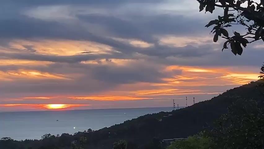 The video shows a sunset over the ocean, viewed from an elevated position with trees in the foreground. The sun is setting on the horizon, with orange and red hues illuminating the sky and reflecting on the water. There are dark gray and light gray clouds streaking across the sky.  The foreground features a verdant, tree-covered hillside. A bird flies across the scene. Communication towers are visible on the hilltop.