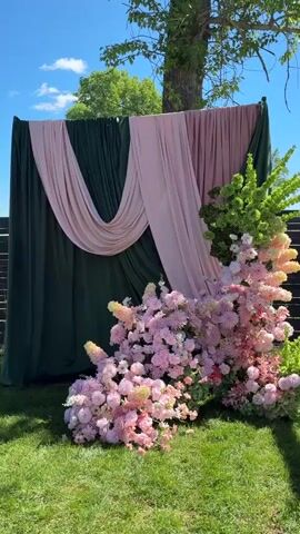 The scene features a backdrop composed of alternating panels of dark green and light pink fabric draped elegantly. In front of the backdrop sits an arrangement of pink flowers, accented by some greenery. The backdrop and floral arrangement are situated on a patch of bright green grass. A tree with green foliage can be seen just behind the backdrop. The sky is blue and partly cloudy.