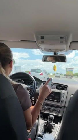 This is a POV shot from the back seat of a car driving on a highway on a sunny day. The driver, a woman wearing sunglasses and a grey t-shirt, is holding a smartphone mounted on the dashboard in her right hand. Her left hand is on the steering wheel. The phone screen is visible and shows some content. A dashboard with air vents, a radio, and gear shift is also visible. A yellow emoji air freshener hangs from the rearview mirror. Outside the car, other vehicles including cars and a truck are visible on the highway. The sky is blue with some clouds. Two disposable coffee cups are in a holder in the center console.