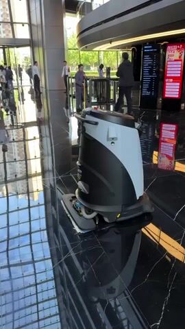 A black and gray robot floor cleaner is parked on a polished black marble floor in a building lobby. The robot is in the center of the frame. People walk around the robot in the background, seemingly ignoring it. To the right, a person stands in front of a digital movie poster. To the left, a person uses a mop. The lobby has shiny floors that reflect the surrounding interior and exterior. The building has a modern design with glass windows and metal accents. The overall atmosphere is busy, with people moving through the lobby.