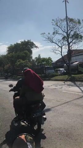 A person riding on the back of a motorcycle, wearing a dark jacket and a maroon hijab. The motorcycle is traveling on a paved road in a semi-urban area. Other vehicles and buildings are visible in the background, along with trees and a clear blue sky. The perspective is from behind the motorcycle, looking forward.