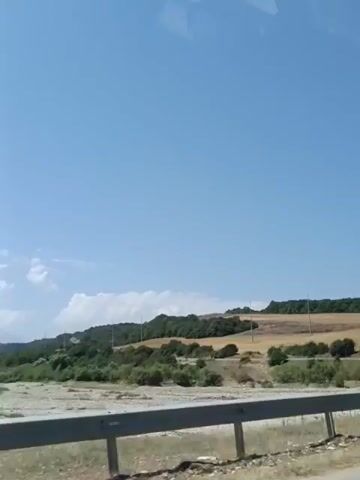 A scenic view of a rural landscape under a clear blue sky. The foreground features a metal guardrail. Beyond the guardrail, there's a dry, rocky riverbed. In the middle ground, scattered green bushes and trees dot the landscape, with a large expanse of dry, golden-brown fields stretching towards a distant tree line. The background is dominated by rolling hills covered in trees and the expansive sky. Power lines are visible on the hills. The overall impression is of a dry, sun-drenched countryside.