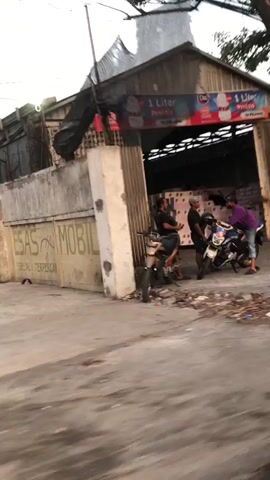 In Gelam, East Java, this media captures a daytime scene outside a building. Three men are present, two seated on motorcycles and one standing, seemingly engaged in a conversation or interaction. One man is wearing a purple hoodie and jeans, while the other two are dressed casually. The motorcycles appear to be parked outside a corrugated metal structure, possibly a shop or workshop, which has a banner above its entrance. The banner displays imagery of ice cream and text, though it's partially obscured and difficult to read entirely. The overall atmosphere appears relaxed, with no strong indicators of specific emotions. The lighting suggests it is daytime, and the weather seems fair. The setting includes a paved area in front of the building, with some debris scattered on the ground, and a large tree trunk partially obstructing the view on the right side of the frame.
