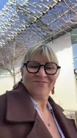 A woman with blonde hair and black-framed glasses is recording a selfie video. She is wearing a brown coat over a blue collared shirt and a silver necklace. Behind her is a modern, open-air structure with a lattice-like roof. A clothing store is visible in the background, featuring mannequins in the display windows. A person wearing a blue jacket walks by in the background. The woman maintains a direct gaze at the camera with a slight smile.