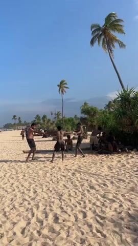 The video opens on a sunny beach with a clear blue sky. In the foreground, a sandy beach is visible, with several palm trees and lush green vegetation in the background. Several people are gathered on the beach. Two men are engaged in what appears to be a playful fight or sport, with others watching and some sitting in the background. The scene then shifts to a wider view of the beach, showing a long stretch of sand with many people walking and playing near the water's edge. The ocean is visible, with waves gently rolling onto the shore. A red flag is planted in the sand in the foreground of the final shot, signaling a potentially dangerous area. The overall atmosphere is one of leisure and recreation on a tropical beach.