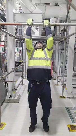 A person wearing a high-visibility vest, hard hat, and gloves is performing a hanging leg raise exercise. They are suspended from a horizontal bar, with their arms fully extended and their legs straight and together. The person is in an indoor industrial or construction environment, with scaffolding and pipes visible in the background. The lighting is bright and uniform.