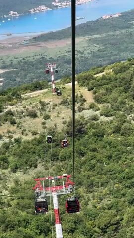 The video opens with a downward view from a cable car, showcasing a series of red and black cable car cabins descending over a lush green, hilly landscape. Some cabins appear to have passengers. One cabin has "Mirac" visible on its side. In the background, a large body of water and a distant town are visible under a bright sky.

The camera then pans upwards and sweeps across a spectacular panoramic vista. A vast, deep blue body of water, resembling a bay or sea, dominates the scene, bordered by numerous green, tree-covered mountains and hills. Along the coastline, a sprawling town or city with densely packed buildings can be seen. A prominent feature is an airport runway, clearly visible on a landmass extending into the water, complete with small aircraft and airport infrastructure. Numerous boats and yachts are scattered across the serene blue water. The sky is clear with a few fluffy white clouds, suggesting a sunny day. The overall impression is one of expansive natural beauty and a vibrant coastal settlement.