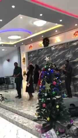 Here's a description of the video:

The video shows a group performing in what appears to be a church or event hall.  The setting includes a decorated stage with a Christmas tree in the foreground.  A man in a dark suit is leading the singing with a microphone, and other singers, mainly women, are backing him up.  A band is also present, with a man playing the drums, a keyboardist, and a guitarist.  Speakers are visible on the side of the stage, and decorations are along the walls. The room has a modern look, with patterned floors and decorated ceiling.