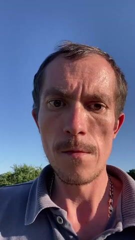A close-up shot focuses on the face of a man with a concerned expression. He has a thin mustache and stubble, and his skin appears flushed, suggesting he may have been exercising or exposed to the sun. The background is a blur of green foliage and a clear blue sky, indicating an outdoor setting. He is wearing a light blue collared shirt and a silver chain.